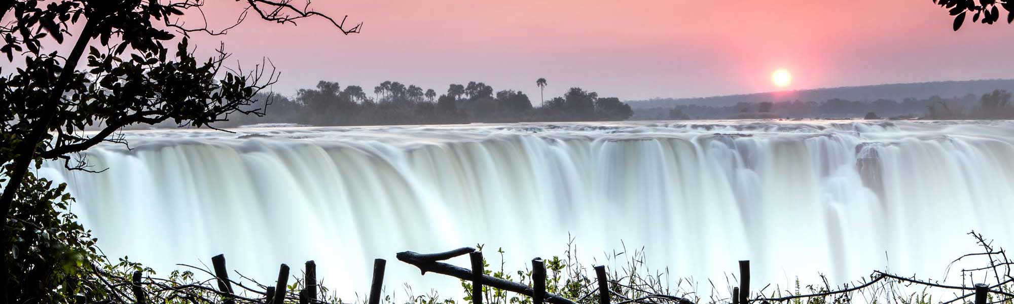 Victoria Falls at Sunrise with Mist A breathtaking view of Victoria Falls at sunrise, with massive cascading water creating a luminous white curtain against a pink dawn sky. The iconic waterfall is framed by lush vegetation and trees along the Zambezi River, with the sun rising majestically in the background.