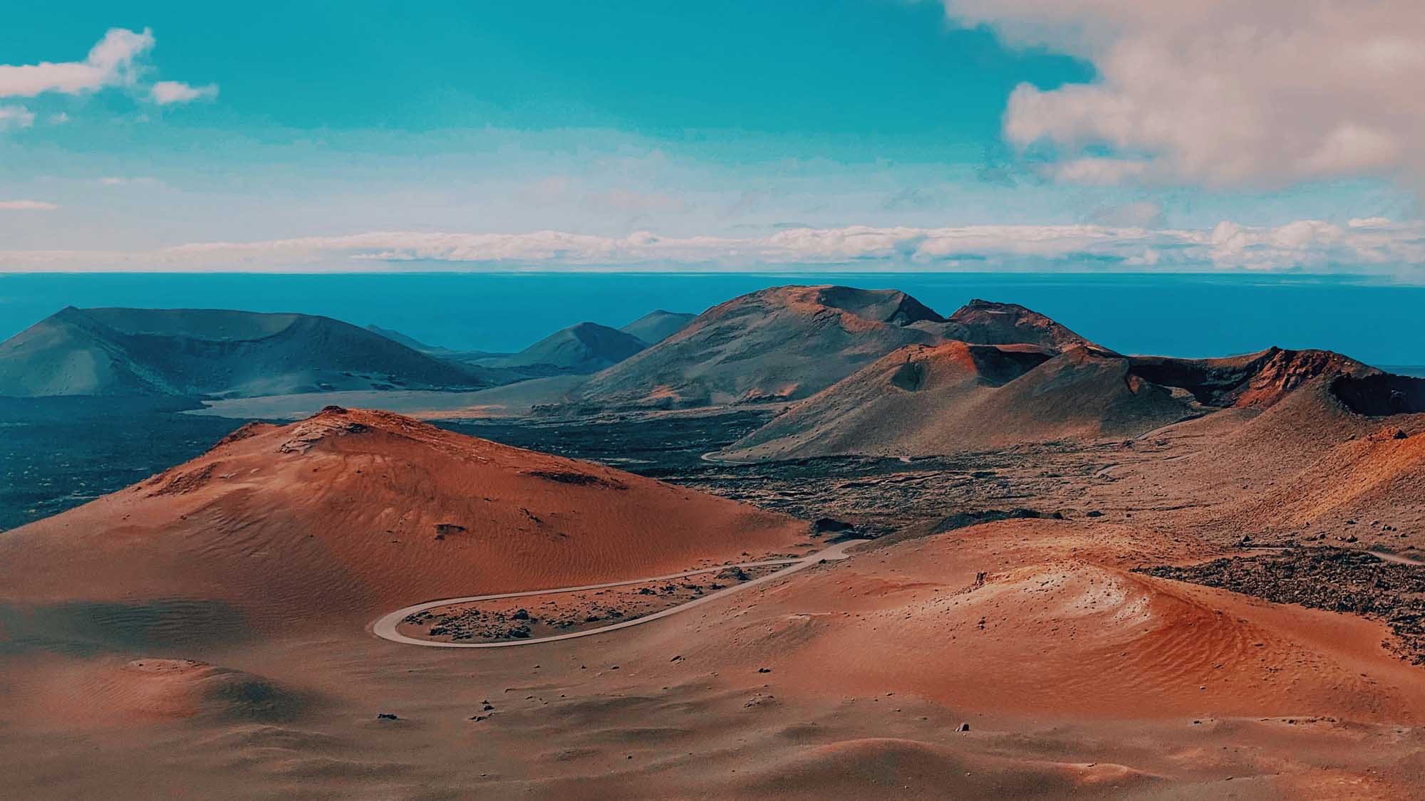 Volcanic Landscape with Turquoise Ocean A dramatic volcanic landscape featuring distinctive rust-red and dark cinder cones rising above a vast lava field, with a striking turquoise ocean and distant islands visible on the horizon. The arid terrain displays the raw geological beauty of a volcanic archipelago with minimal vegetation.