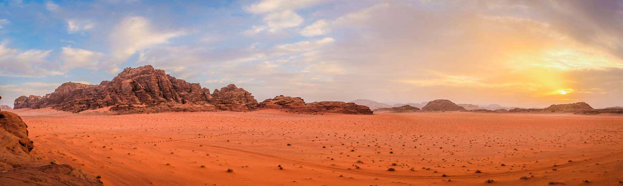 Wadi Rum Desert Landscape at Sunset A breathtaking panoramic view of the Wadi Rum desert in Jordan, featuring towering rust-colored sandstone rock formations rising from the expansive red sand plains during golden hour, with a dramatic sky transitioning from blue to warm orange and yellow tones.