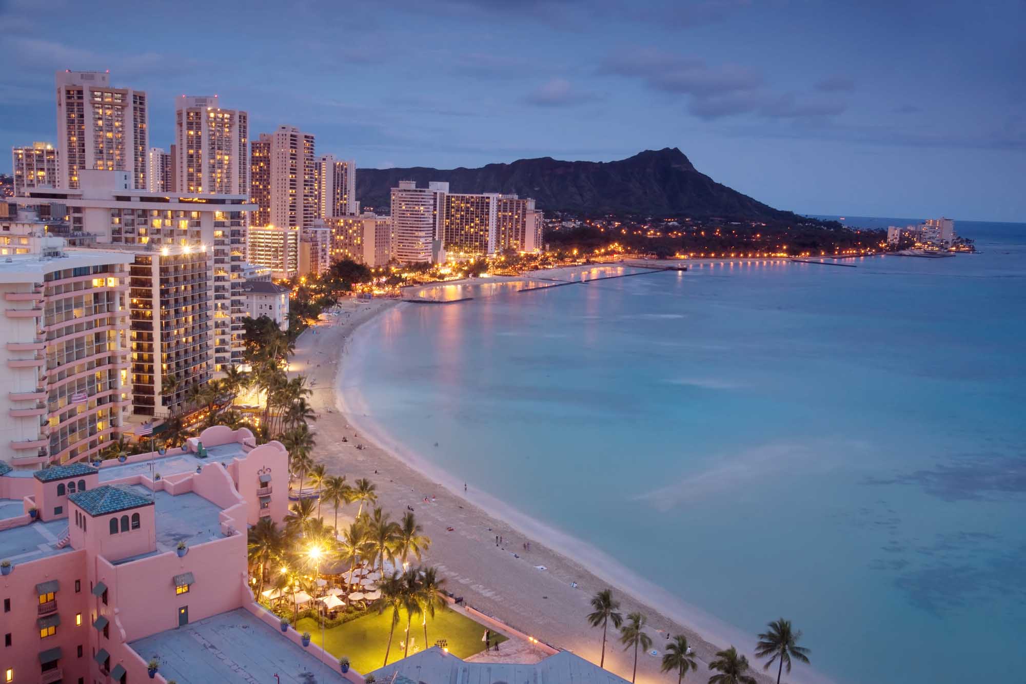 Waikiki Beach at Twilight with Diamond Head A stunning twilight view of Waikiki Beach, Hawaii, showcasing the iconic crescent shoreline lined with luxury high-rise hotels and resorts illuminated against the deepening blue sky. Diamond Head volcanic crater dominates the background, while palm-lined beaches and calm turquoise waters reflect the warm glow of evening lights along the bustling beachfront.