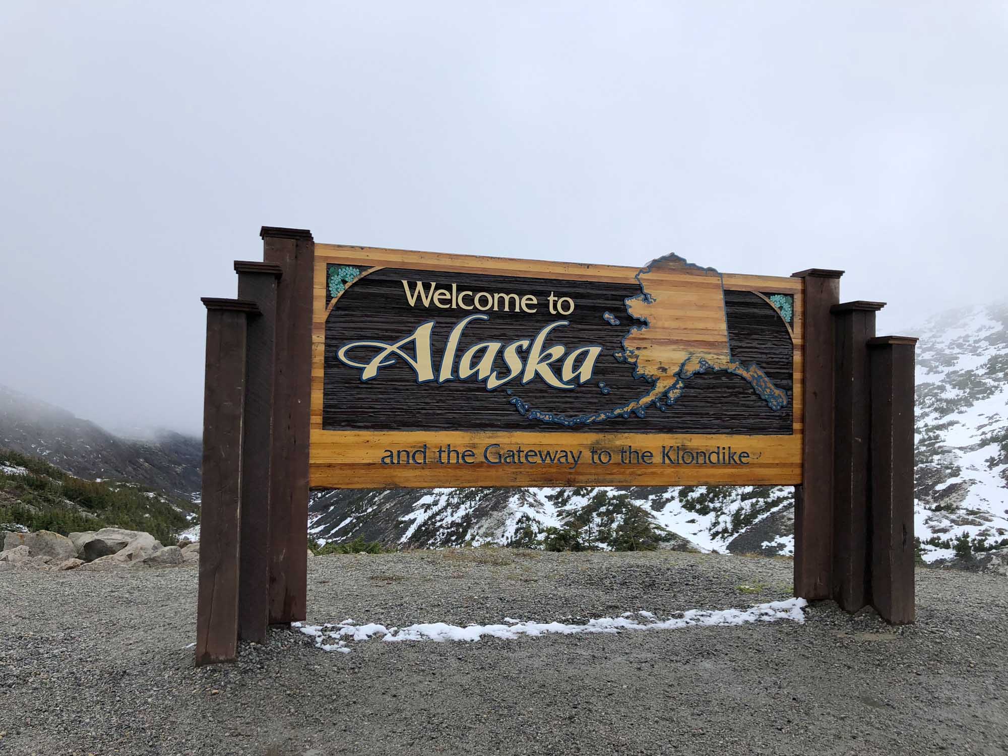 Welcome to Alaska Gateway to Klondike Sign A rustic wooden welcome sign marking the entrance to Alaska, identifying it as the gateway to the Klondike. The sign features carved text and the state outline against a backdrop of snow-capped mountains and misty valleys.