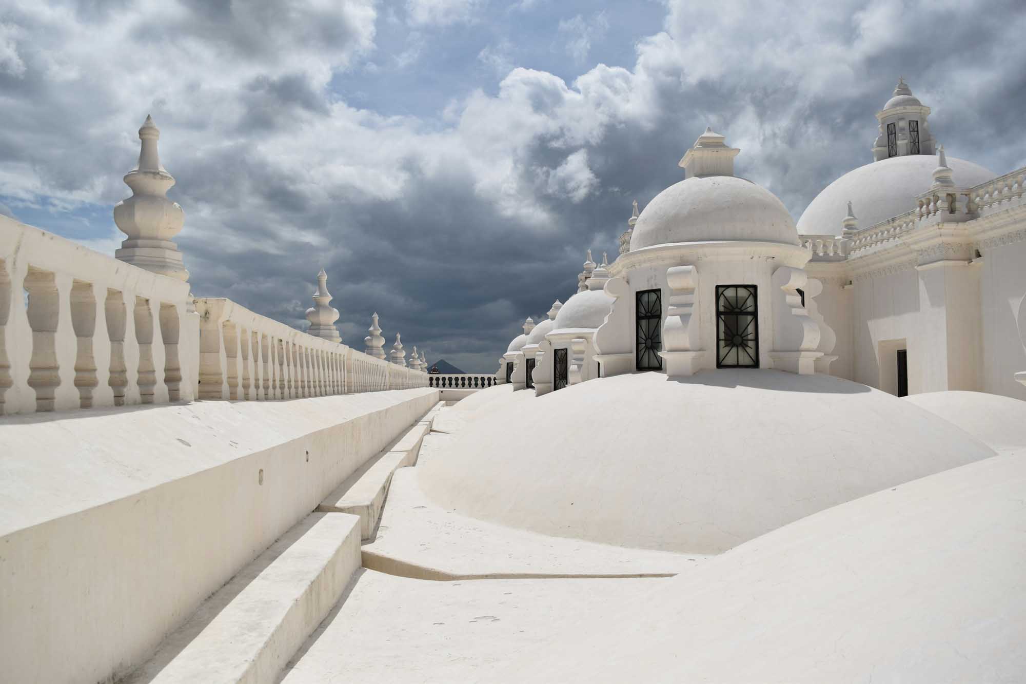 White Domed Architecture Rooftop Terrace A stunning rooftop terrace featuring pristine white-washed architecture with iconic domed structures and ornamental balustrades, characteristic of Mediterranean or Middle Eastern design. The dramatic cloudy sky creates a striking contrast against the brilliant white buildings adorned with decorative finials and geometric details.