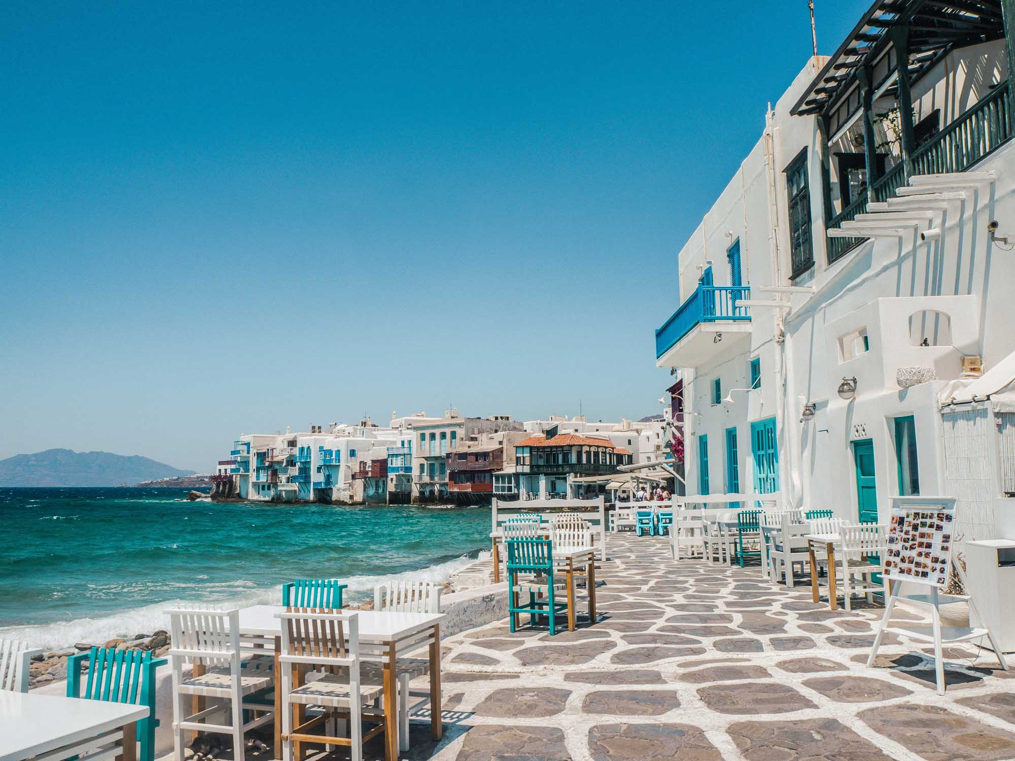 Whitewashed Waterfront Taverna in Little Venice A picturesque Greek island waterfront scene featuring whitewashed buildings with turquoise accents and wooden chairs arranged on a traditional patterned stone plaza overlooking crystal-clear turquoise waters. Charming architecture typical of the Cycladic islands lines the waterfront, creating an idyllic Mediterranean coastal setting.