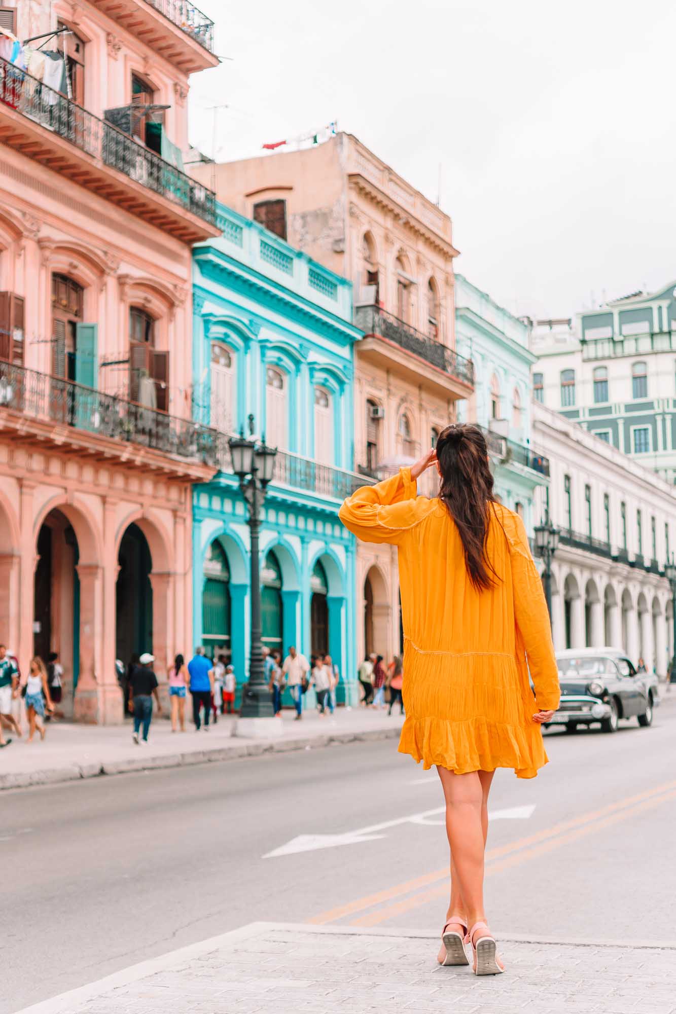 Woman in Yellow Dress, Old Havana Street A young woman wearing a vibrant yellow dress stands on a colorful street in Old Havana, Cuba, gazing at the beautifully preserved colonial architecture featuring pastel pink, turquoise, and cream-colored buildings with ornate balconies and arched windows.