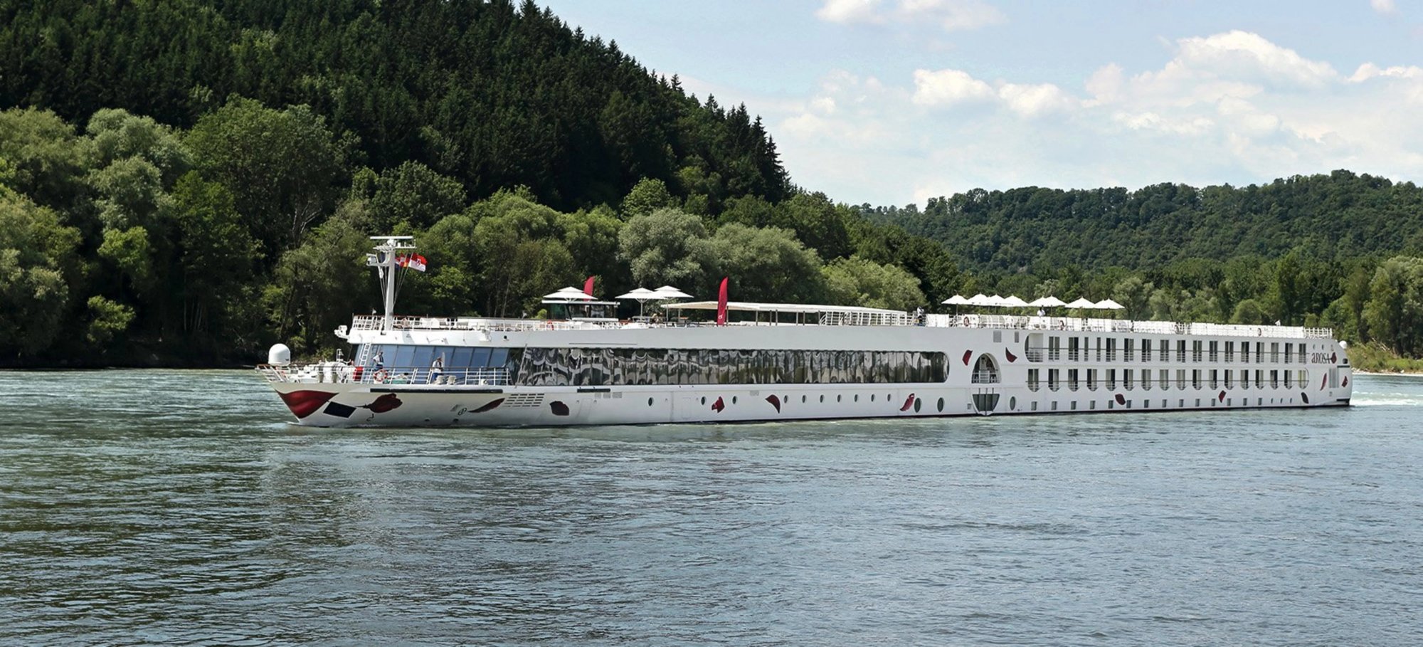 A modern river cruise ship operated by A-ROSA navigates a scenic waterway lined with dense forests