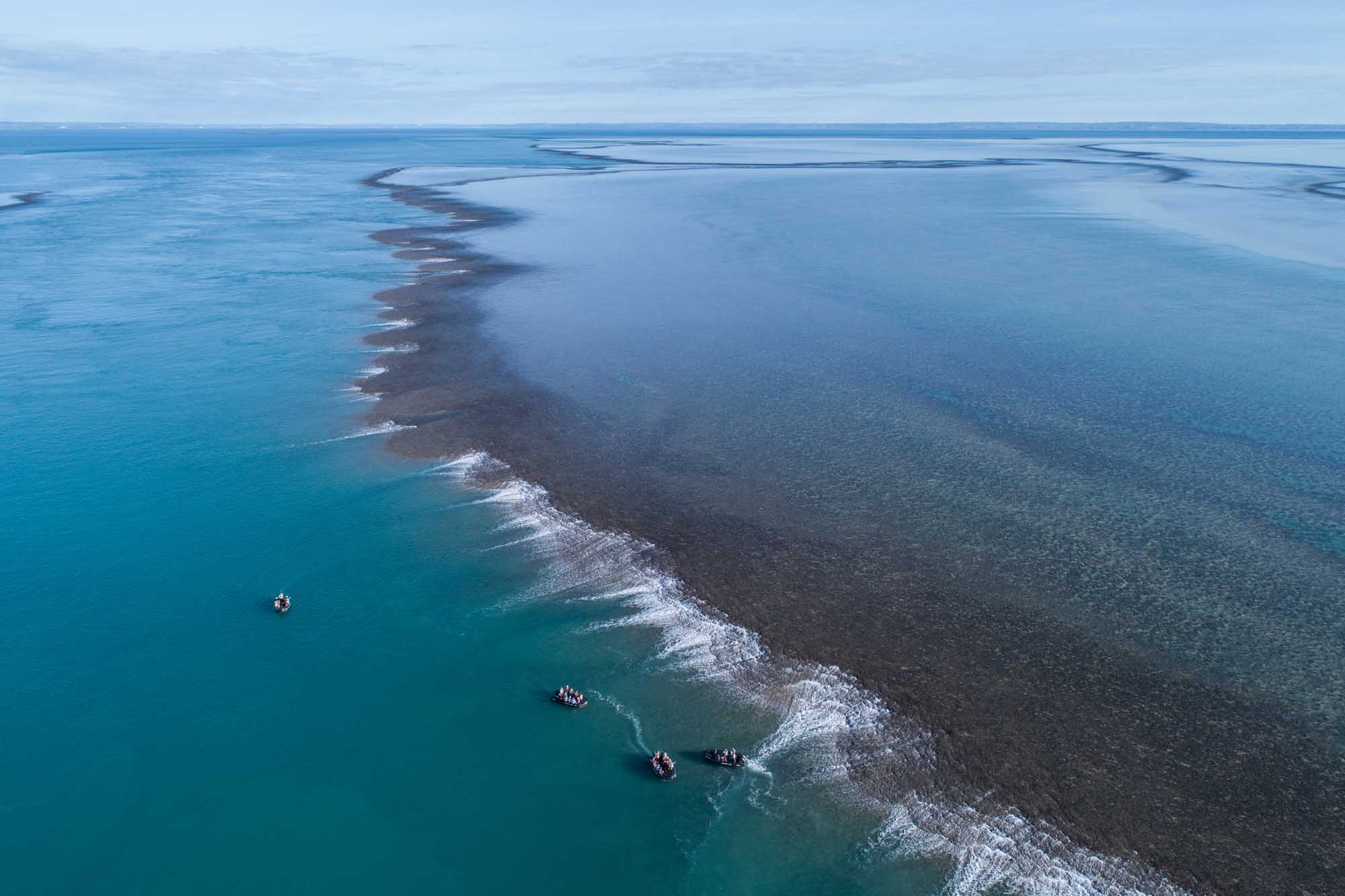 Aerial View of Coastal Reef Formation and Boats An aerial perspective of a dramatic coastal landscape featuring a distinctive dark reef or sandbank