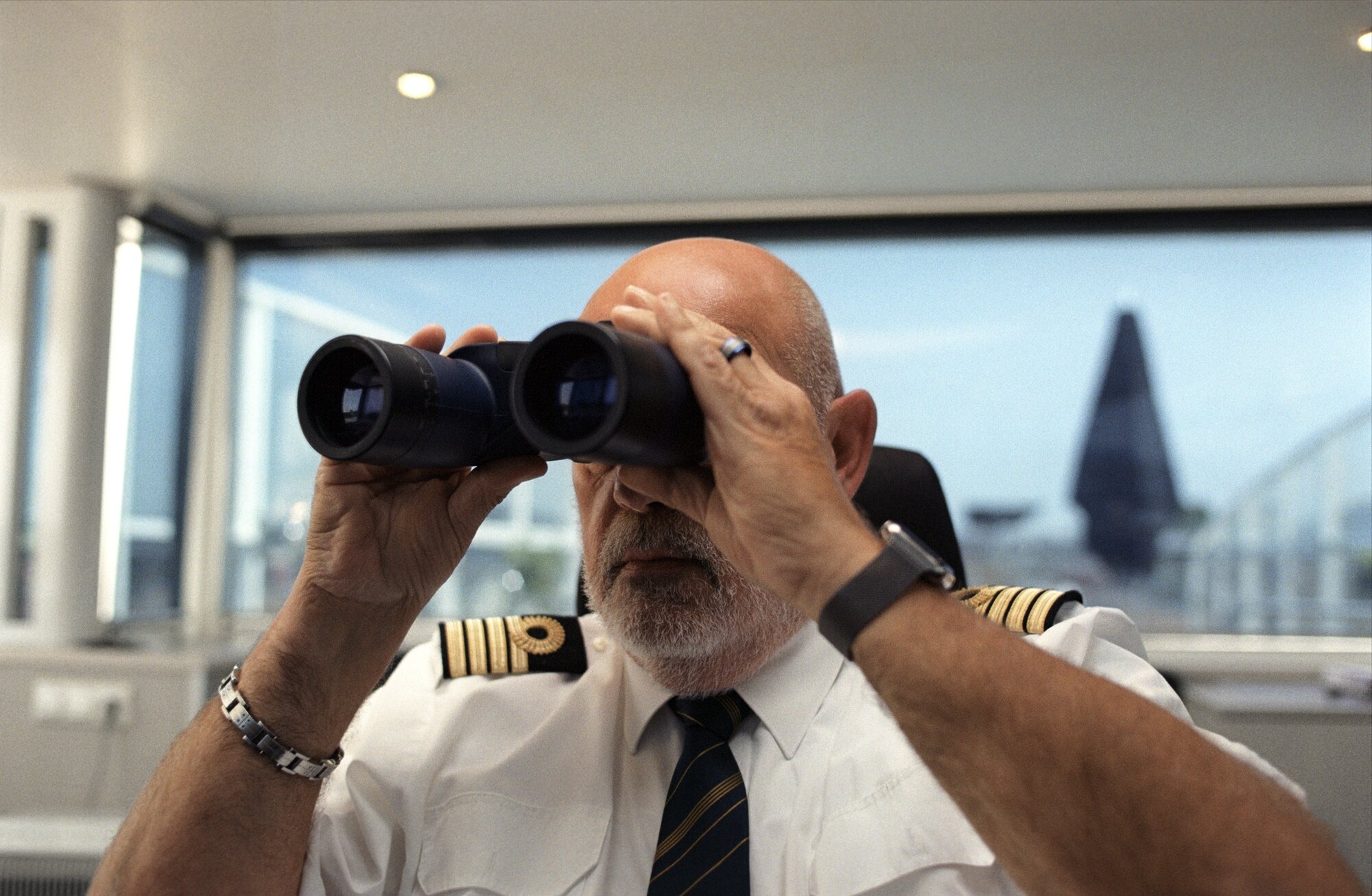 Captain Observing from Ship Bridge with Binoculars A senior captain in white uniform shirt and black tie, wearing officer's insignia on his sleeves