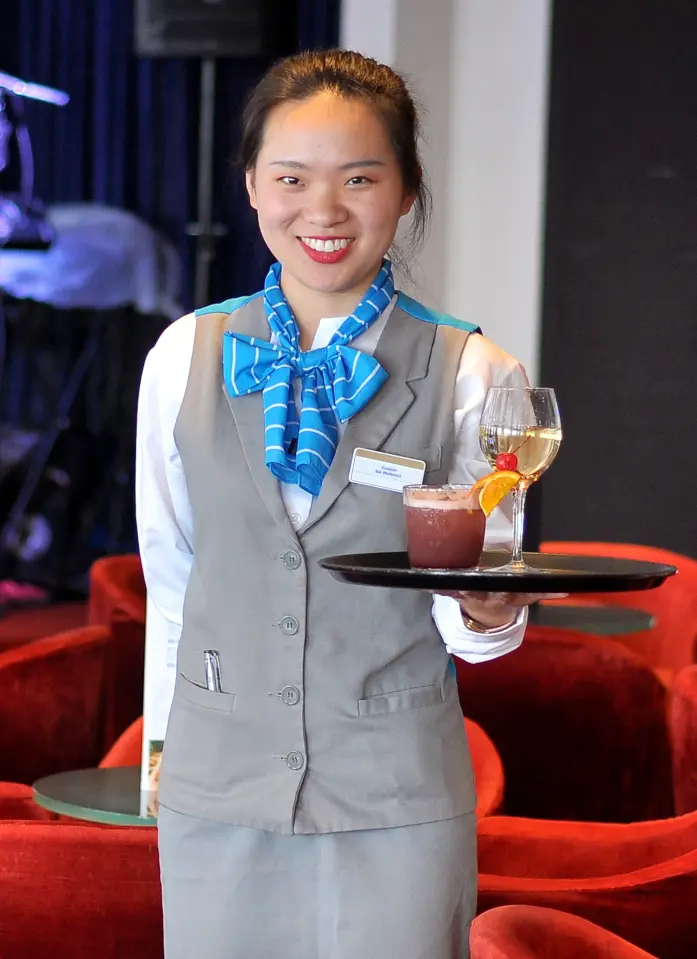 Celestyal Cruises Crew Member with Beverages A smiling crew member in Celestyal Cruises uniform (gray vest with blue scarf) stands in an elegant