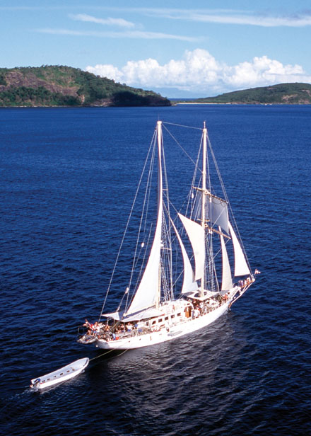 A stunning white-sailed tall ship navigates the brilliant blue waters of Fiji, with lush, forested