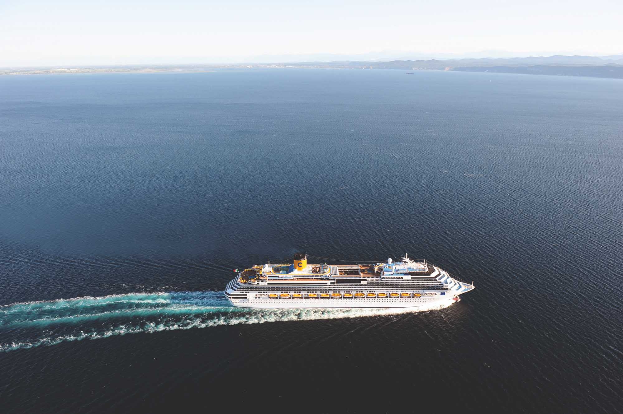 An aerial view of a large Costa cruise ship sailing through calm blue waters, with distinctive