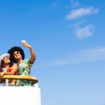 A couple stands on an upper deck of a cruise ship against a bright blue sky, with one person