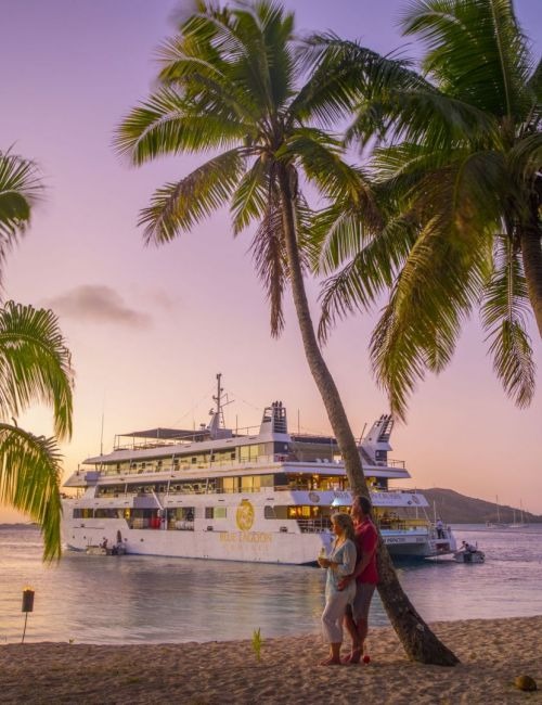 A luxury cruise ship anchored in calm tropical waters at sunset, photographed from a pristine beach