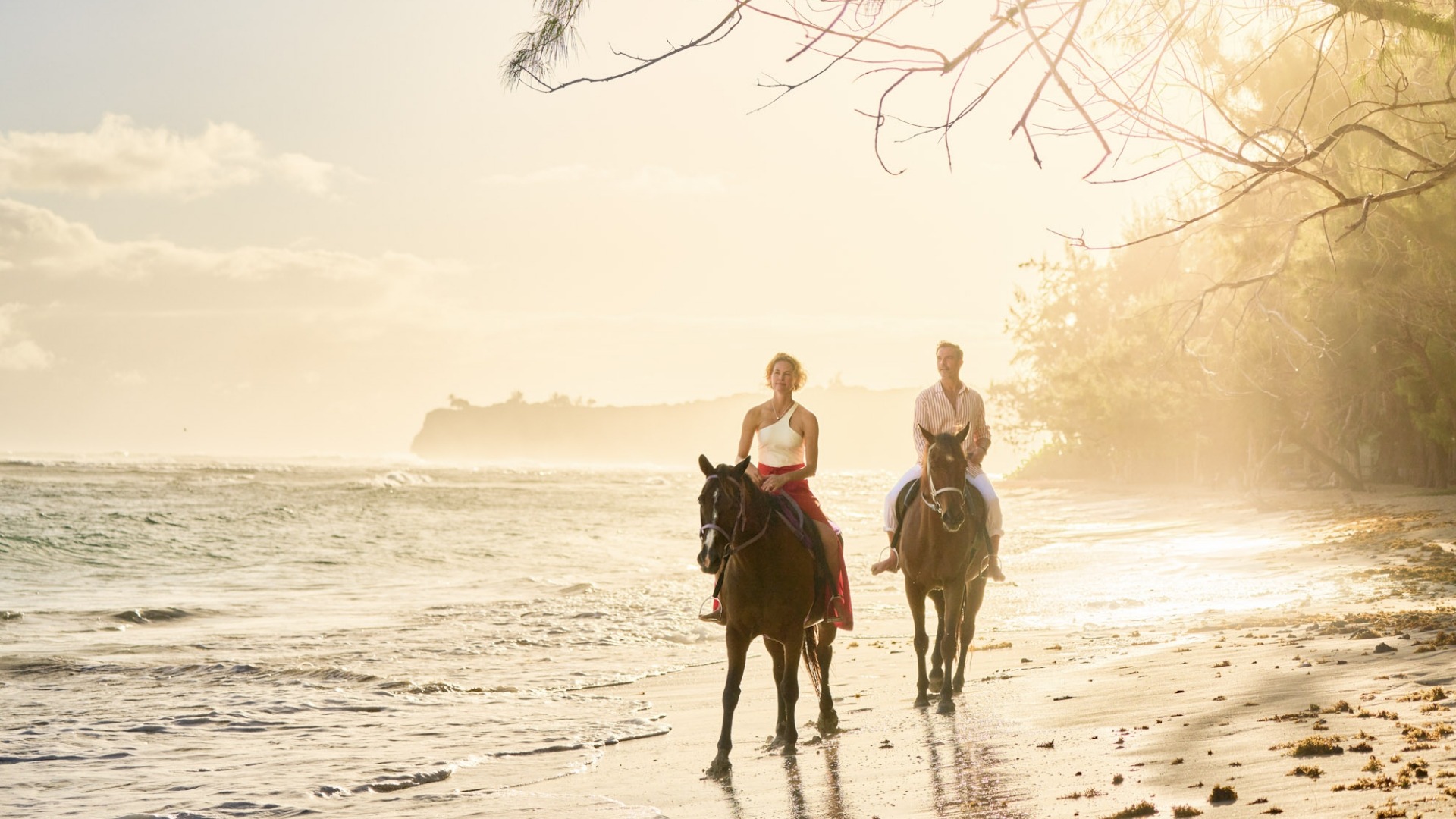 Horseback Riding on Tropical Beach at Sunset Two riders on horseback traverse a pristine sandy beach at golden hour, with gentle waves lapping
