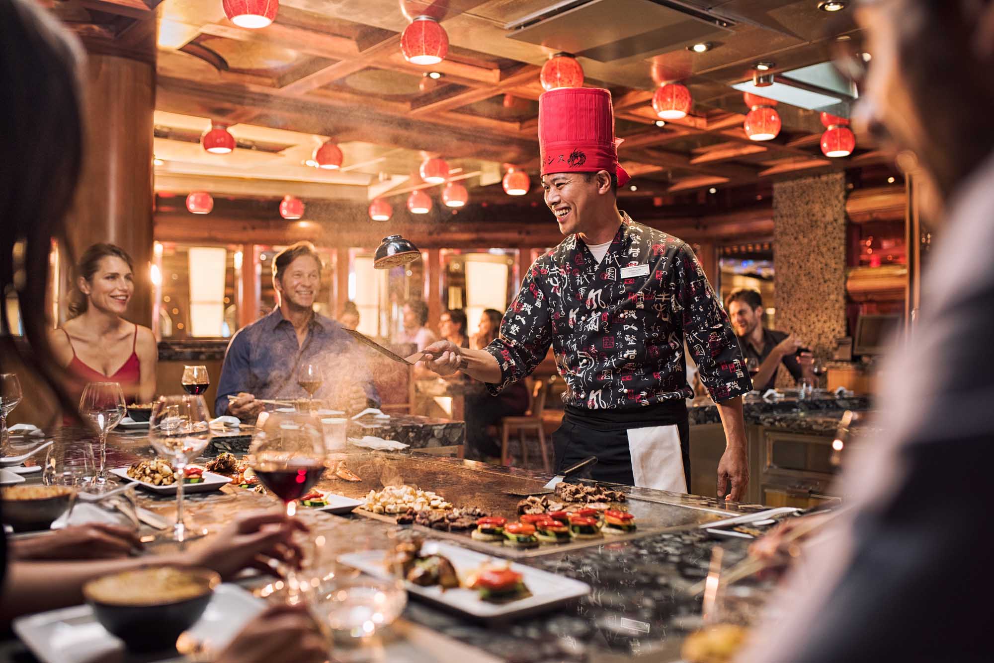 Interactive Teppanyaki Dining Experience on Cruise Ship A chef in traditional red hat and decorative black jacket prepares food at an open teppanyaki