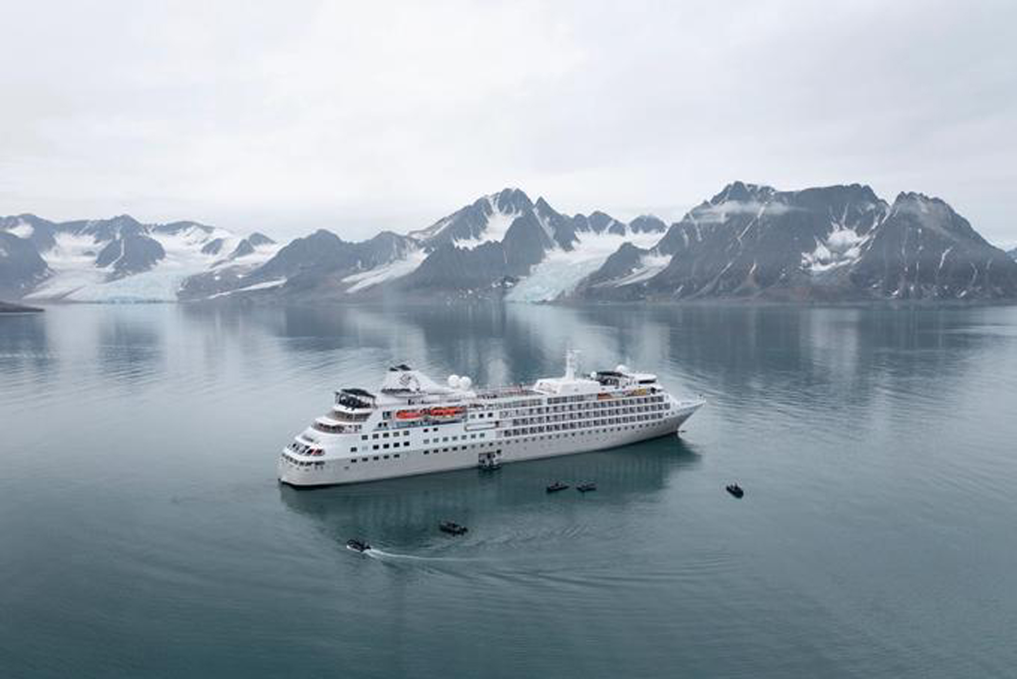 Luxury Cruise Ship in Arctic Waters with Glacial Mountains A large white expedition cruise ship anchors in calm, mirror-like Arctic waters with small tender