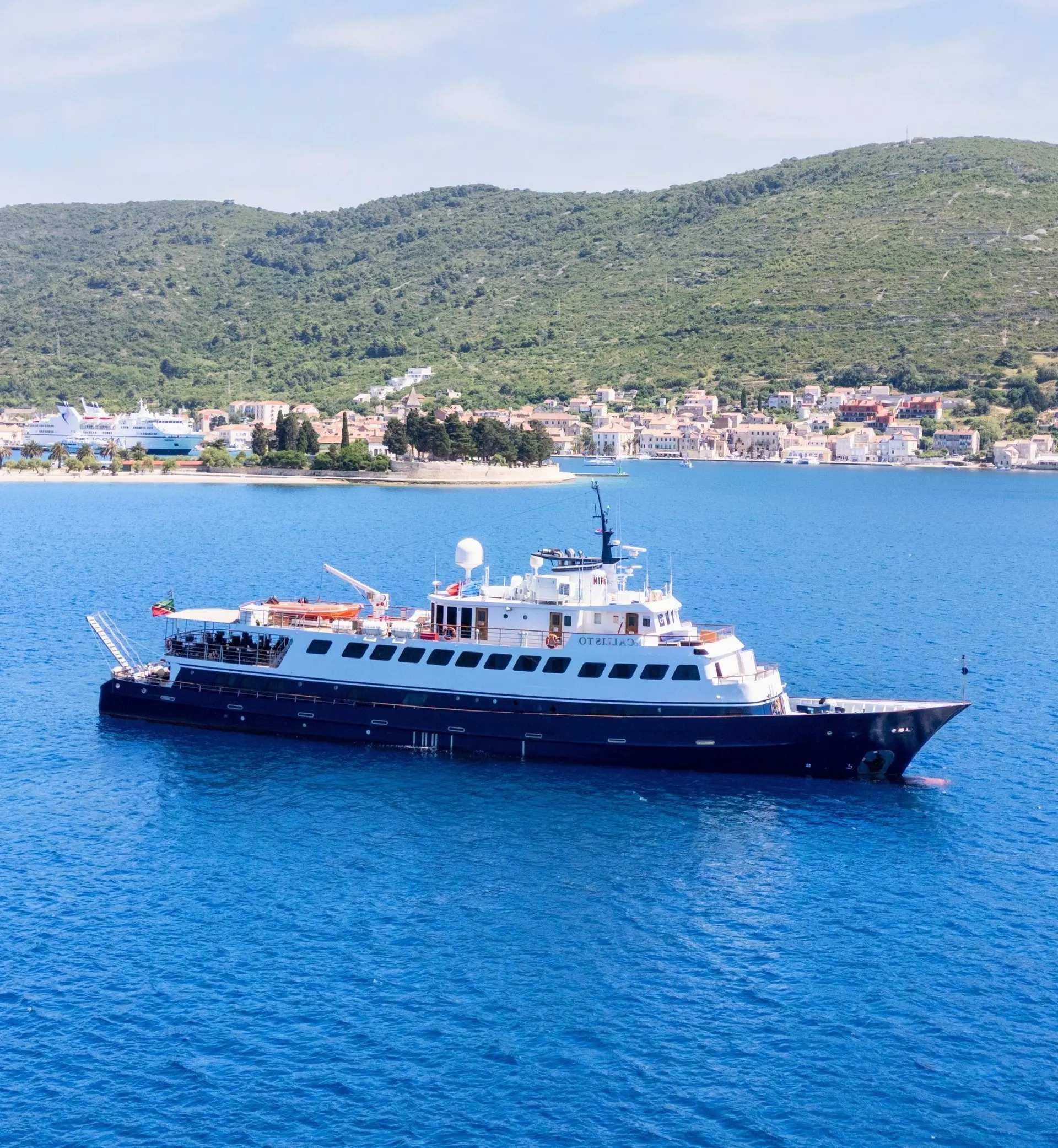 A sleek expedition cruise ship with dark blue and white hull anchored in crystal-clear turquoise