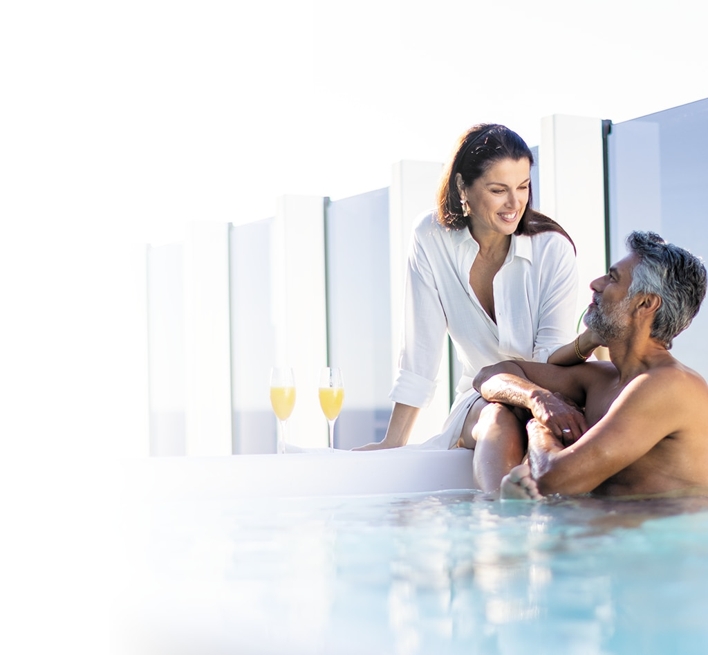 A woman in a white shirt serves champagne to a man relaxing in a sunlit hot tub or jacuzzi