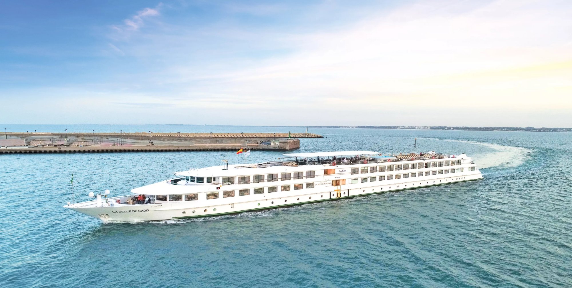 A modern river cruise ship, MS La Belle de Cadix, anchored in turquoise waters near a port facility