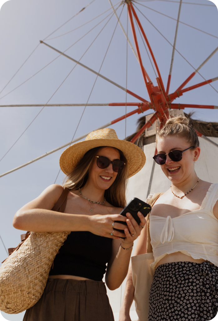 Passengers Enjoying Celestyal Cruise Deck Two smiling women on a cruise ship deck, wearing sunglasses and summer attire, looking at a mobile