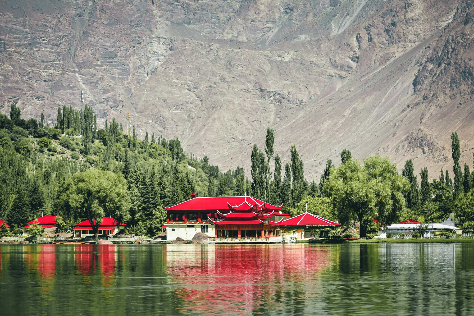 Red-Roofed Temple by Mountain Lake in Skardu A striking traditional Asian temple with distinctive red tiered roofs is reflected in the calm