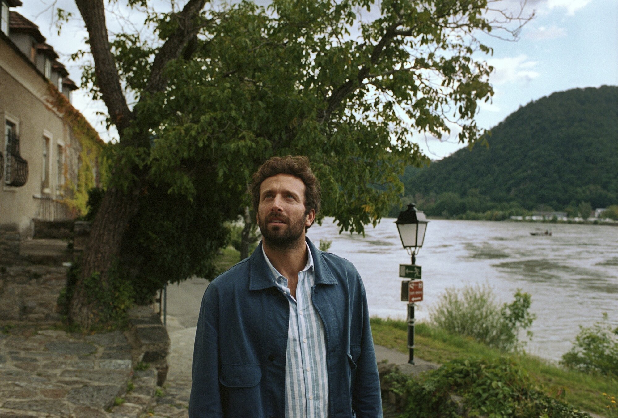 Solo Traveler at Riverside Village in European Valley A bearded man in casual attire stands along a picturesque riverside promenade beneath weeping