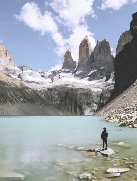 Turquoise Lake with Granite Towers Mountain Landscape A solitary hiker stands on rocky shore beside a pristine turquoise glacial lake, gazing up