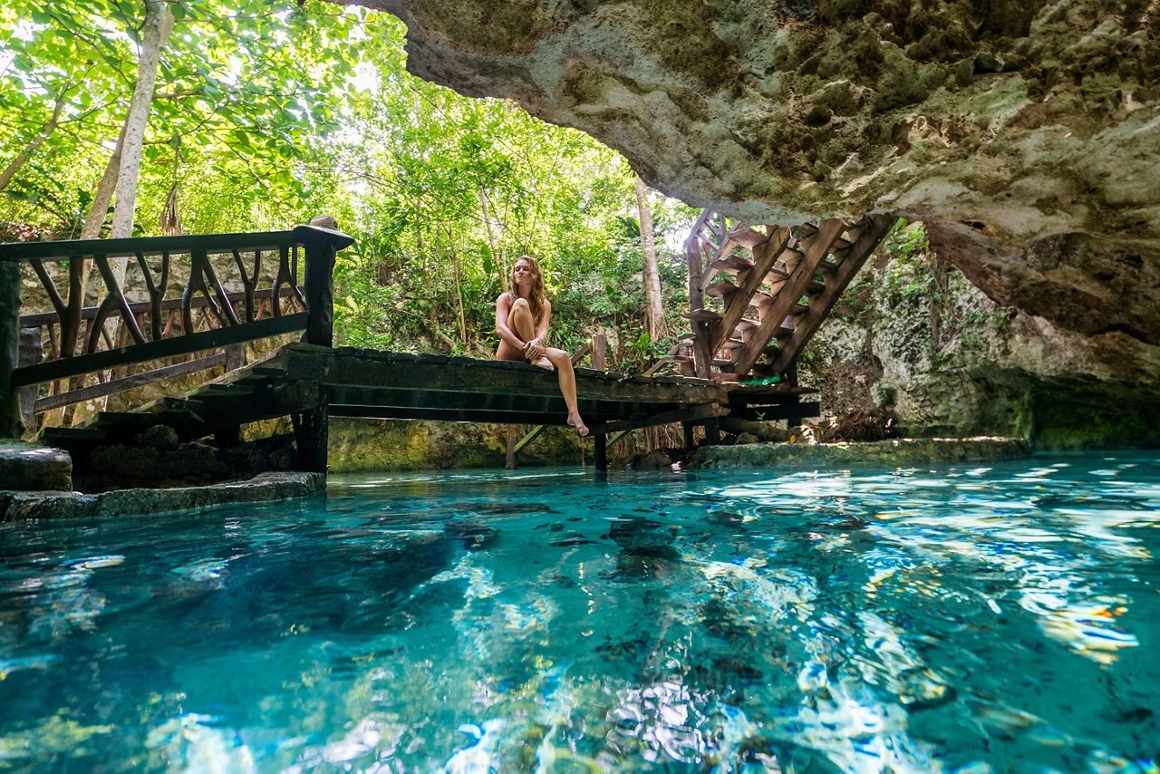 A person sits on a weathered wooden dock extending over crystal-clear turquoise waters beneath