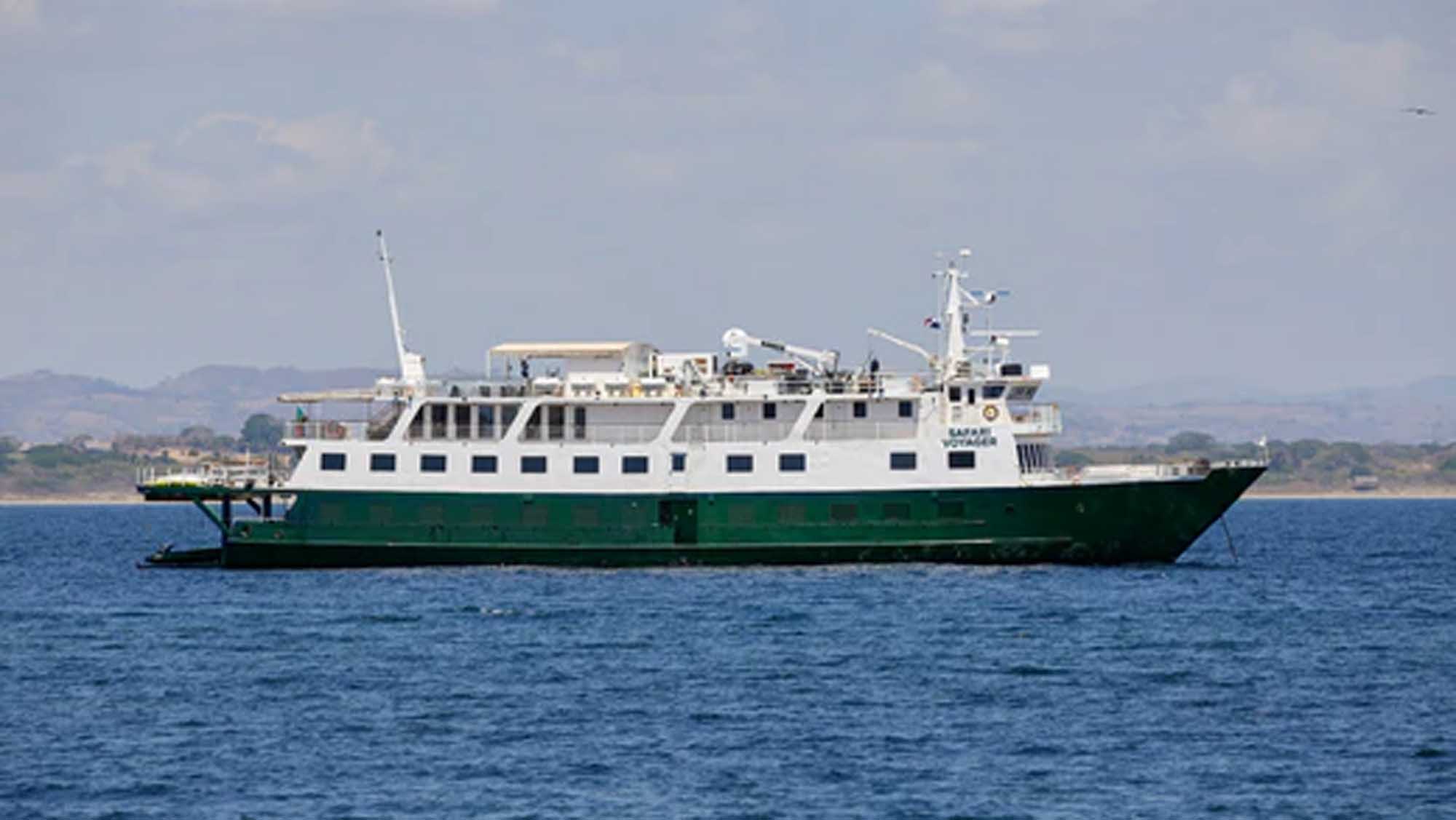 A white and green expedition cruise ship anchored in calm blue waters with a coastal landscape